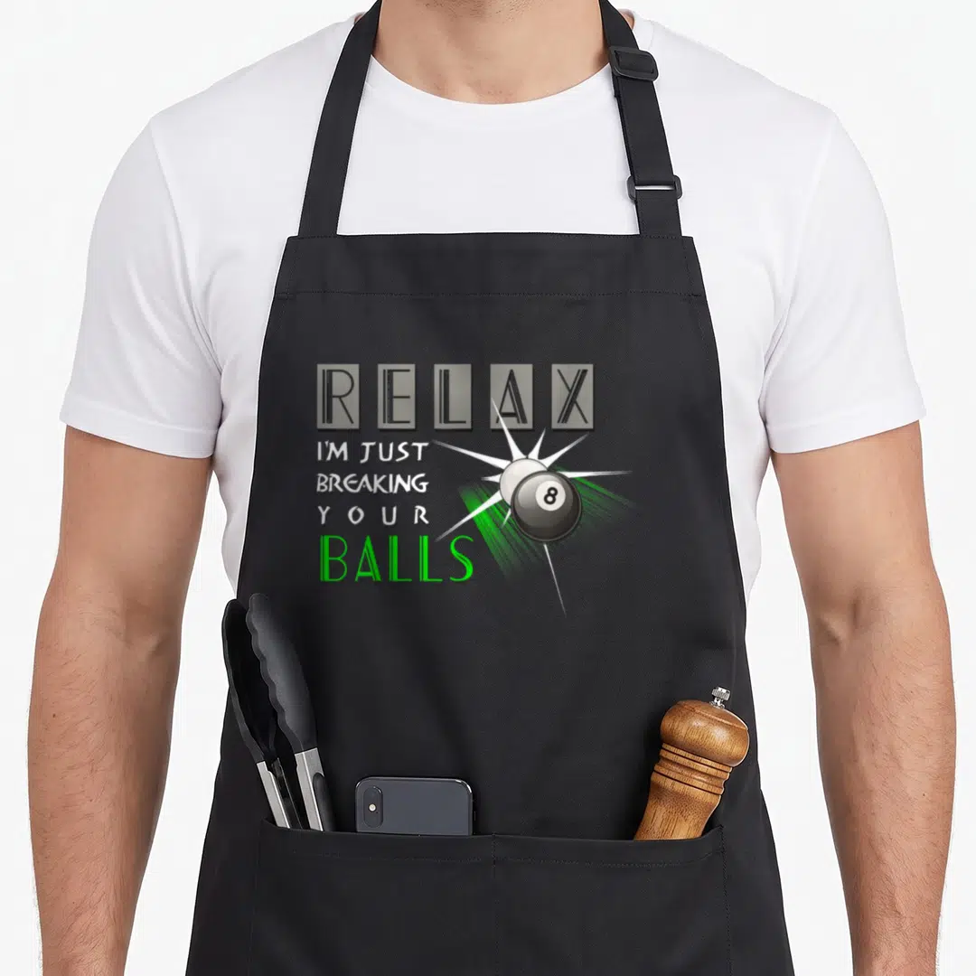 Breaking billiards lover apron - RELAX I'M JUST BREAKING YOUR BALLS text next to cartoon 8-ball collision in green — shown on a black cotton canvas apron against a white background