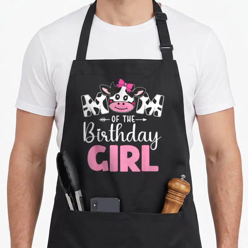 Birthday 1st cow print apron - Cartoon cow head with pink bow above spotted MOM text and pink GIRL lettering — shown on a black cotton canvas apron against a white background