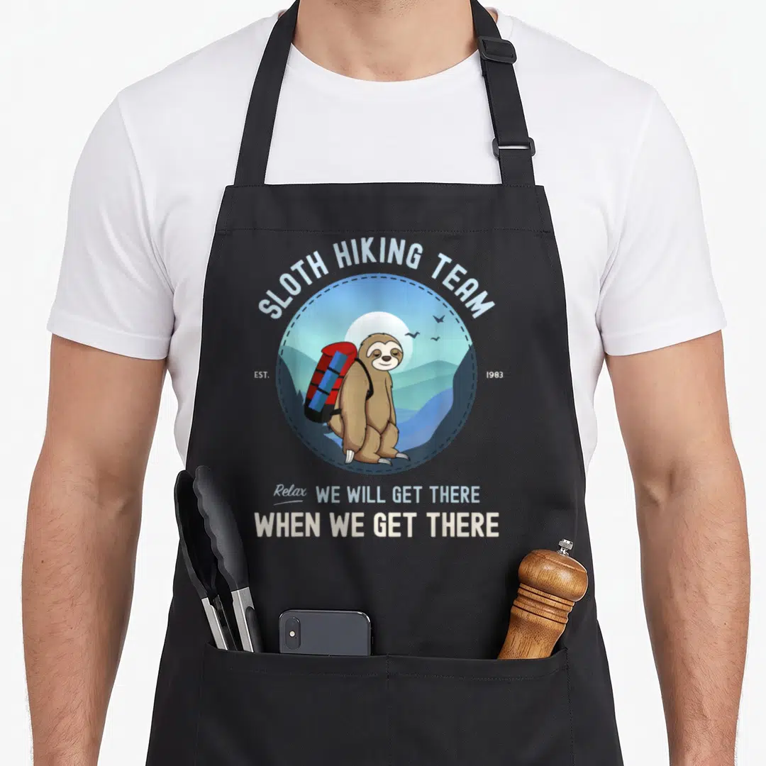 Retro hiking sloth apron - Cartoon brown sloth with red blue backpack inside circle with hiking team text — shown on a black cotton canvas apron against a white background