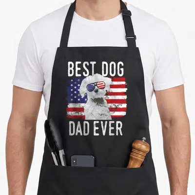 Dog maltese american flag apron - White dog with flag sunglasses between BEST DOG and DAD EVER text on distressed flag — shown on a black cotton canvas apron against a white background