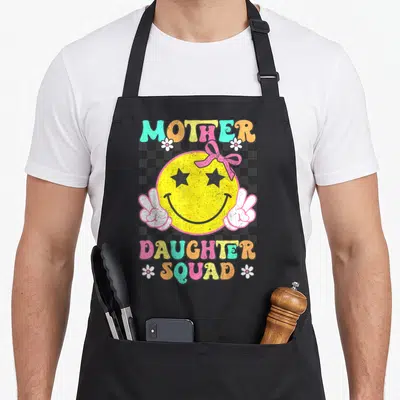 Squad my mother's apron - Retro yellow smiley face with star eyes and peace signs surrounded by MOTHER DAUGHTER SQUAD — shown on a black cotton canvas apron against a white background