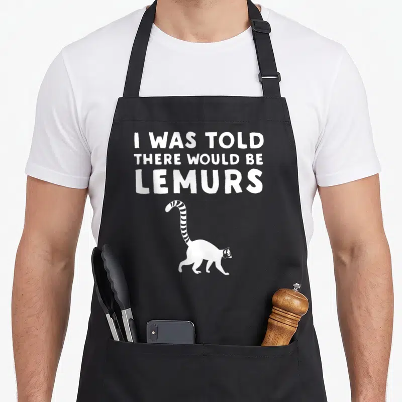 Would lemur lover apron - White text I WAS TOLD THERE WOULD BE LEMURS above a walking cartoon lemur silhouette — shown on a black cotton canvas apron against a white background