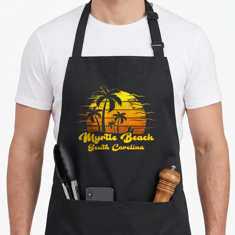 Myrtle tree apron - Retro yellow orange sunset with palm trees and flying birds above Myrtle Beach text — shown on a black cotton canvas apron against a white background