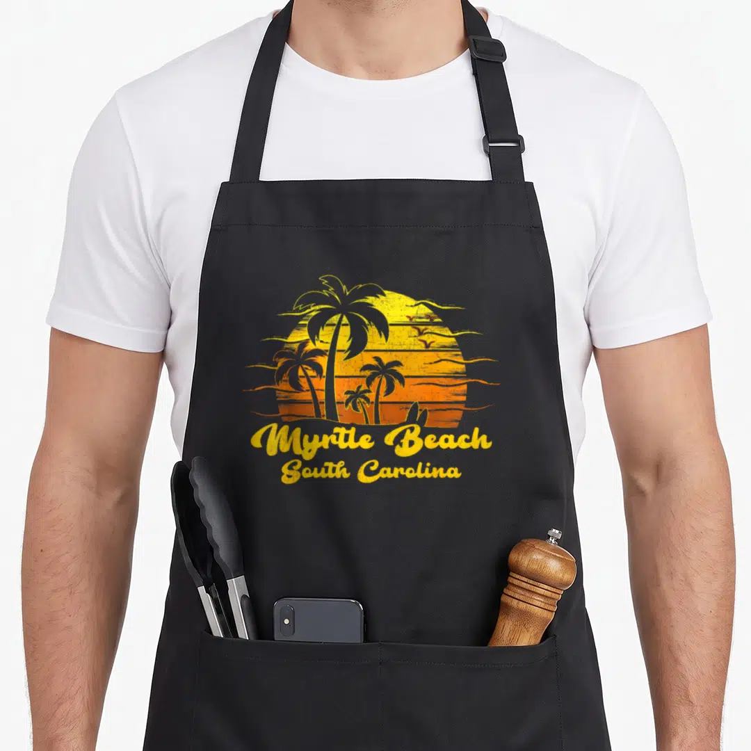 Myrtle tree apron - Retro yellow orange sunset with palm trees and flying birds above Myrtle Beach text — shown on a black cotton canvas apron against a white background