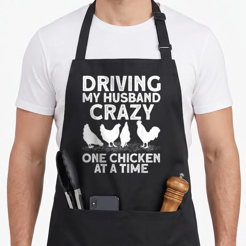 Farmer chicken apron gift - White distressed text Driving My Husband Crazy One Chicken At A Time with four standing — shown on a black cotton canvas apron against a white background