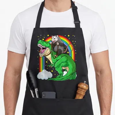 Possum dinosaur apron - Cartoon grey opossum riding green T-Rex with bridle under rainbow and stars — shown on a black cotton canvas apron against a white background