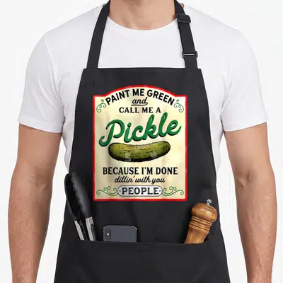 Call pickle apron - Retro sign style graphic with green pickle and text Paint Me Green Call Me A Pickle — shown on a black cotton canvas apron against a white background