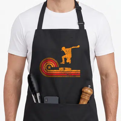 Skateboarder 1970s apron - Orange jumping skateboarder silhouette with red concentric star circles and horizontal — shown on a black cotton canvas apron against a white background