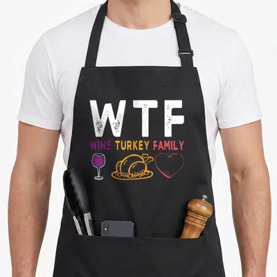 Wine thanksgiving aprons - WTF WINE TURKEY FAMILY text with wine glass, turkey, and heart icons in white purple orange — shown on a black cotton canvas apron against a white background