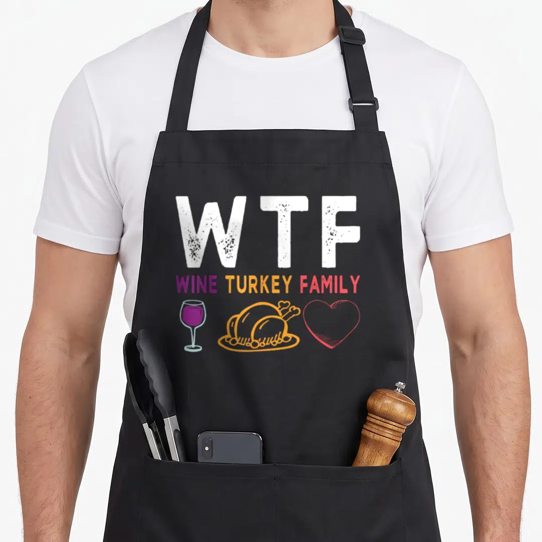 Wine thanksgiving aprons - WTF WINE TURKEY FAMILY text with wine glass, turkey, and heart icons in white purple orange — shown on a black cotton canvas apron against a white background
