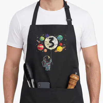 Astronaut 3rd birthday apron - Distressed astronaut holding number 3 moon balloon with planets and rocket in blue white — shown on a black cotton canvas apron against a white background