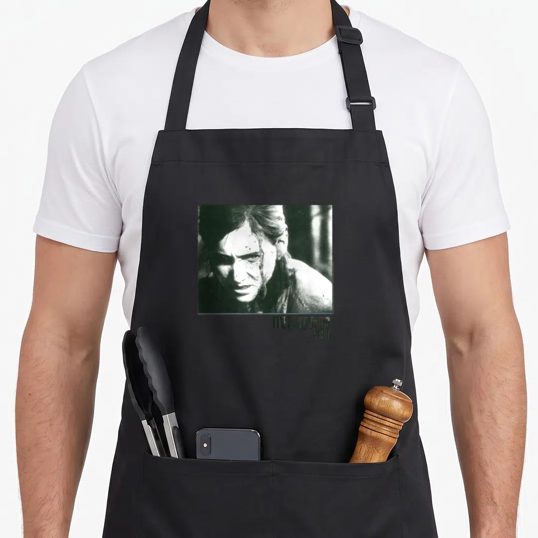Boxed us aprons - Black and white photo of a woman face with THE LAST OF US PART II text below — shown on a black cotton canvas apron against a white background
