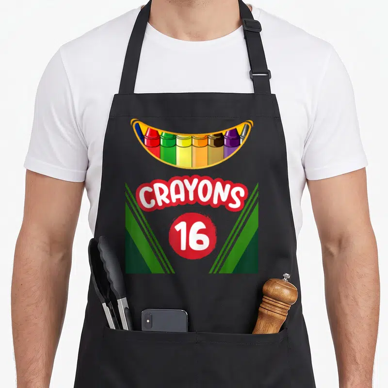 Box teacher apron - Cartoon crayon box graphic with red bubble text CRAYONS and number 16 flanked by green stripes — shown on a black cotton canvas apron against a white background