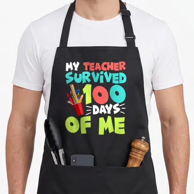 School preschool boys apron - Stacked text My Teacher Survived 100 Days of Me with pencil cup and supplies — shown on a black cotton canvas apron against a white background