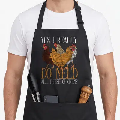 Farm do i need an apron - Three distressed chickens in orange and brown with text Yes I Really Do Need All These — shown on a black cotton canvas apron against a white background