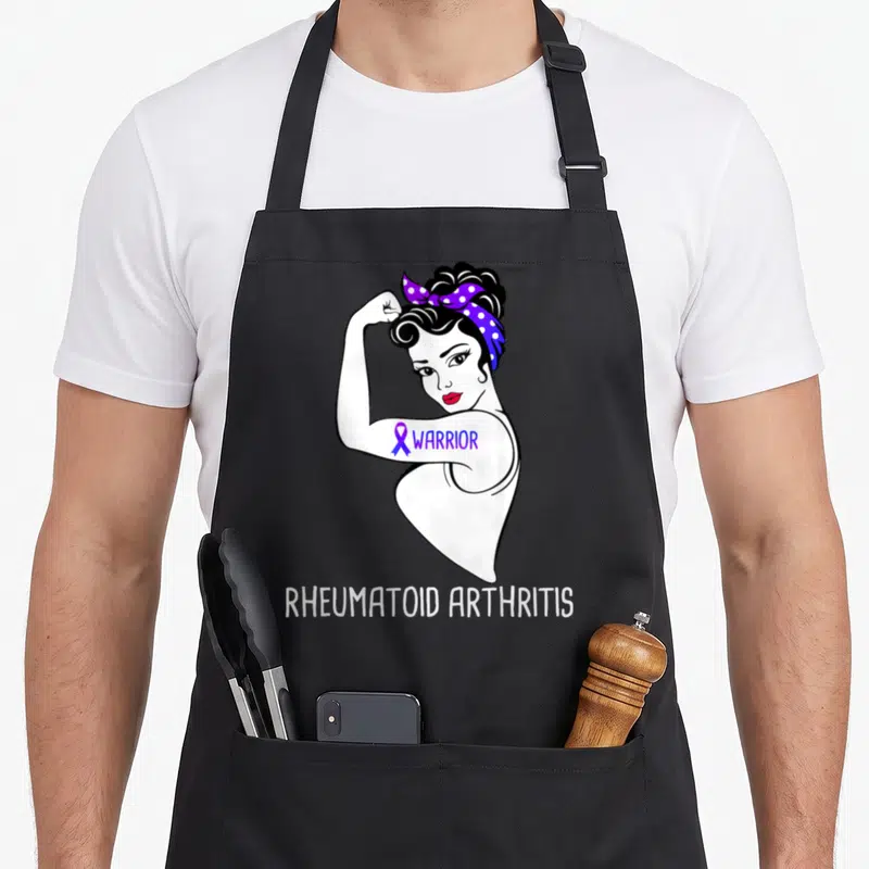 Arthritis ribbon apron - Line art woman flexing arm with purple ribbon and Warrior text below — shown on a black cotton canvas apron against a white background