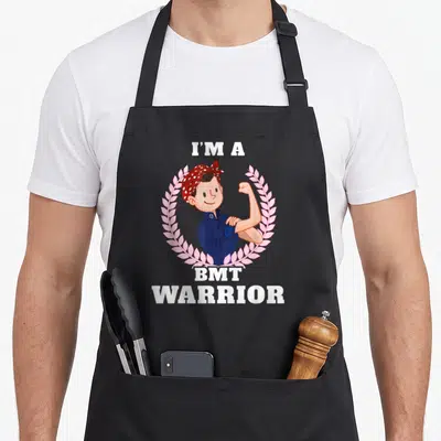Bmt bone cancer awareness apron - Cartoon person flexing arm inside pink wreath with I'm a BMT Warrior text — shown on a black cotton canvas apron against a white background