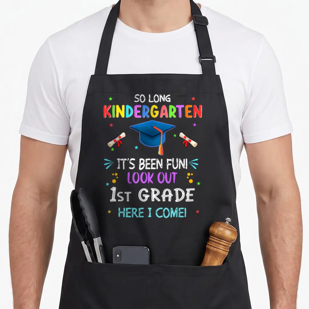 Kindergarten grad apron - So Long Kindergarten text with blue cap, two red-ribboned diplomas, and scattered stars — shown on a black cotton canvas apron against a white background