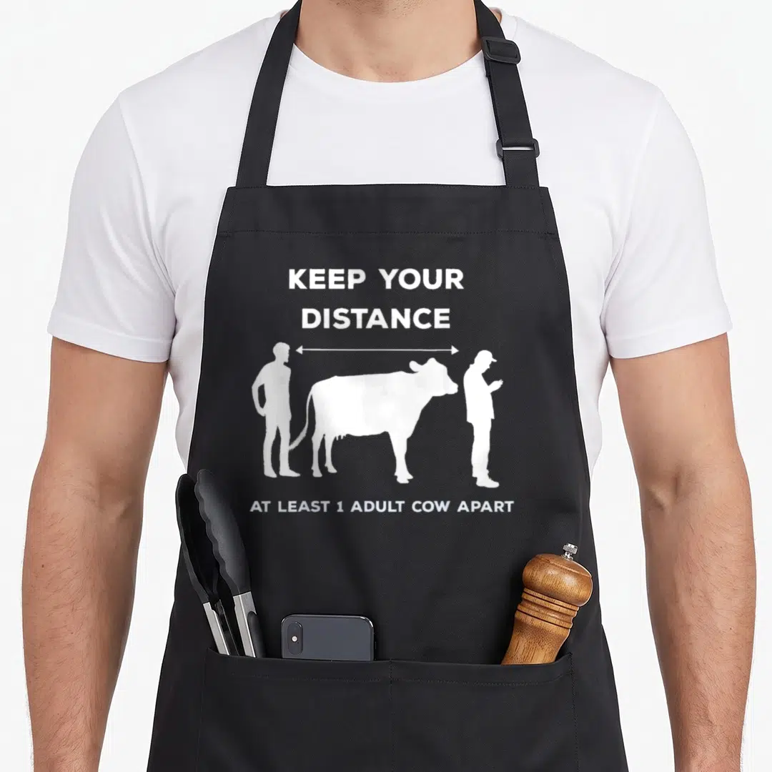 Distance cow apron - White cow and person silhouettes with distance arrow and text on black apron — shown on a black cotton canvas apron against a white background
