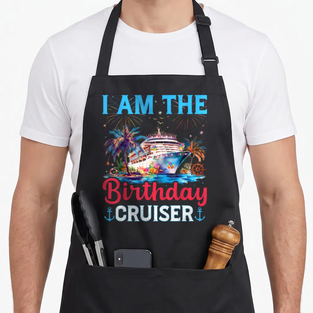 Cruiser birthday apron - Watercolor cruise ship with palm trees, fireworks, lifebuoy, ship wheel, and anchors flanking — shown on a black cotton canvas apron against a white background
