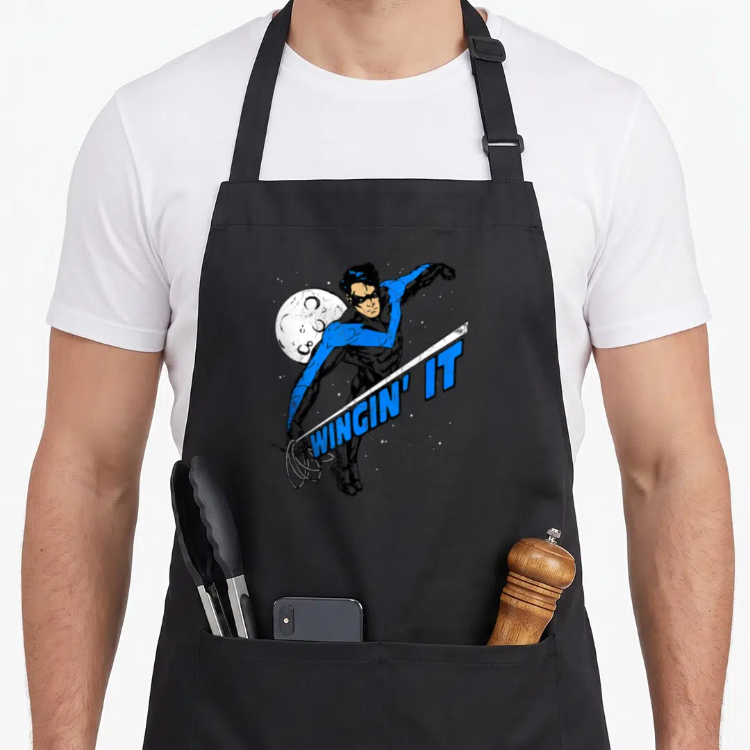 Nightwing machine apron - Nightwing character with moon and diagonal WINGIN' IT text bar — shown on a black cotton canvas apron against a white background