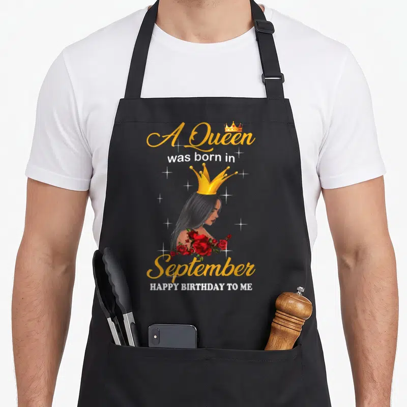 Cute september baking queen apron - Crowned woman with red roses and text A Queen was born in September Happy Birthday — shown on a black cotton canvas apron against a white background