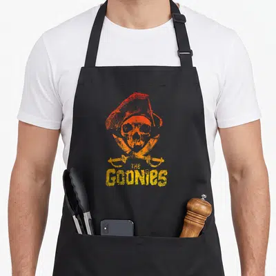 Ombre skull apron - Distressed orange and yellow skull with pirate hat and crossed swords above The Goonies text — shown on a black cotton canvas apron against a white background