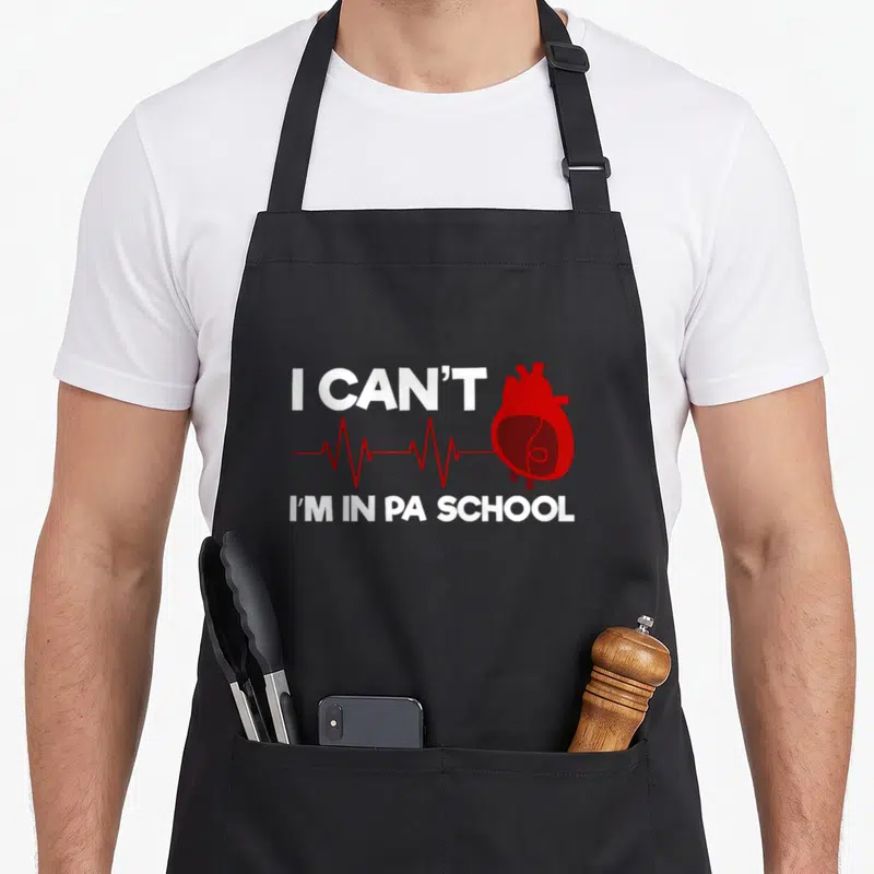 Assistant school physician apron - Red heart and EKG line between white text reading I CAN'T and I M IN PA SCHOOL — shown on a black cotton canvas apron against a white background