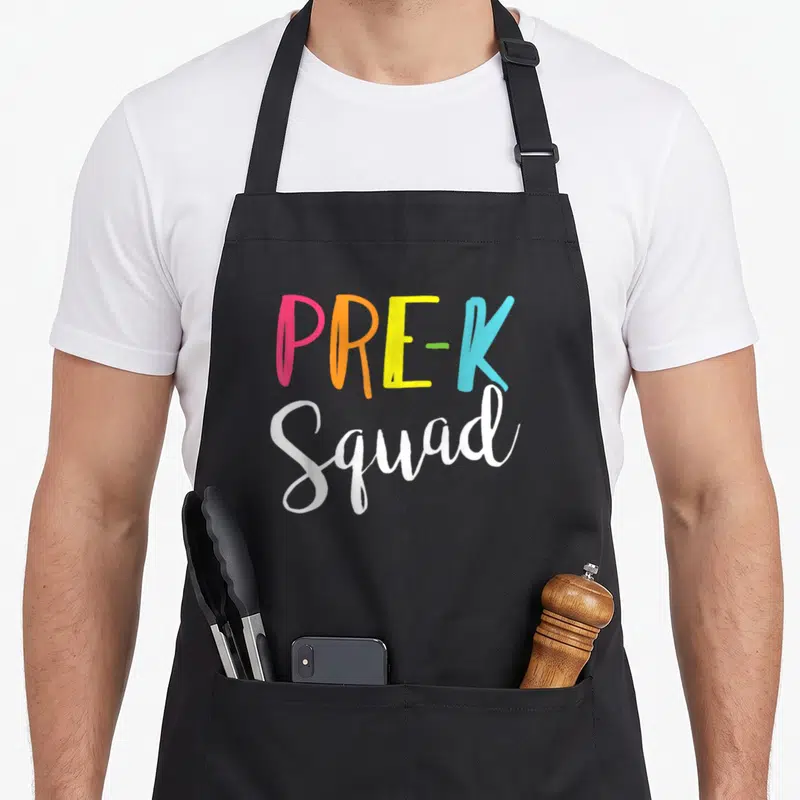 Pre school cute teacher aprons - PRE-K Squad text in pink orange yellow blue and white typography — shown on a black cotton canvas apron against a white background