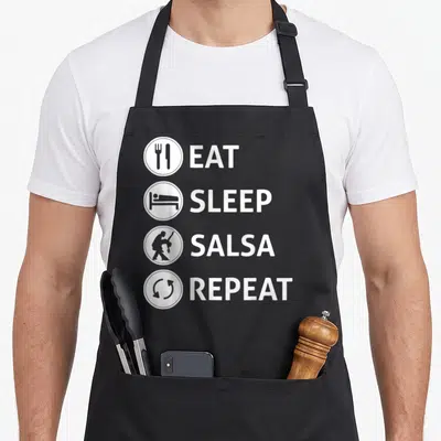 Eat ballroom addict apron - Four white circles with icons next to EAT SLEEP SALSA REPEAT text on black fabric — shown on a black cotton canvas apron against a white background