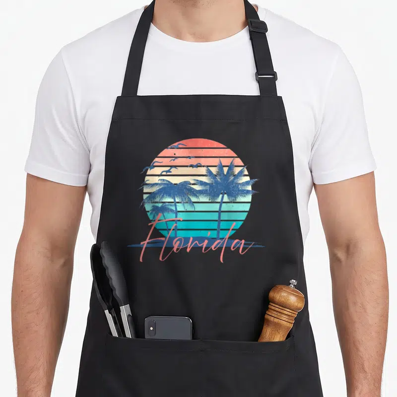 Florida summer tree apron - Retro striped sunset circle with palm tree silhouettes and pink Florida script text — shown on a black cotton canvas apron against a white background