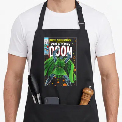 Heroe doctor apron - Retro Doctor Doom comic cover print with green cape and orange spheres — shown on a black cotton canvas apron against a white background