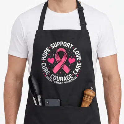 Cancer support ribbon apron - Pink ribbon centered between two hearts with circular text reading HOPE SUPPORT LOVE CURE — shown on a black cotton canvas apron against a white background