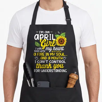 Sunflower april apron - Stacked text reading I'm an April Girl with a yellow sunflower and green leaves flanking — shown on a black cotton canvas apron against a white background