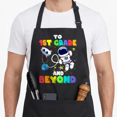 Beyond school apron - Cartoon astronaut rocket and planet with TO 1ST GRADE AND BEYOND text in rainbow colors — shown on a black cotton canvas apron against a white background