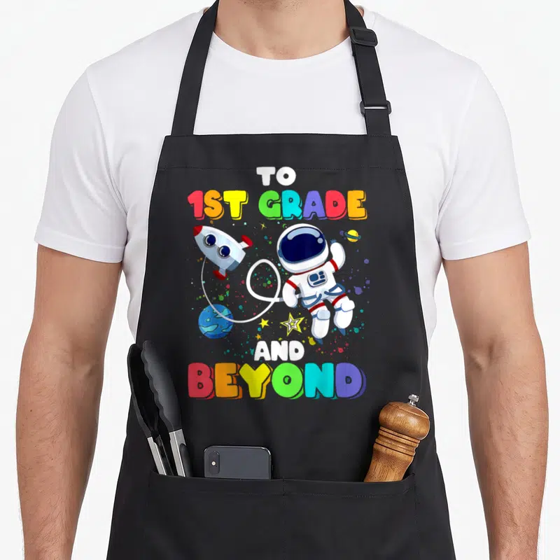Beyond school apron - Cartoon astronaut rocket and planet with TO 1ST GRADE AND BEYOND text in rainbow colors — shown on a black cotton canvas apron against a white background