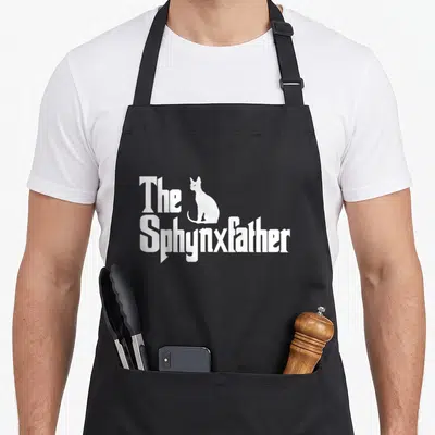 Sphynxfather cat apron - White Sphynx cat silhouette between text The Sphynxfather on black background — shown on a black cotton canvas apron against a white background