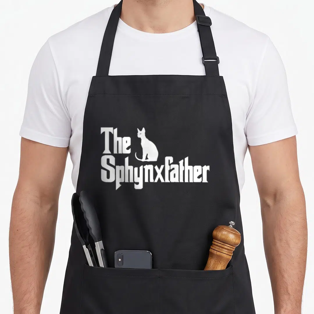 Sphynxfather cat apron - White Sphynx cat silhouette between text The Sphynxfather on black background — shown on a black cotton canvas apron against a white background