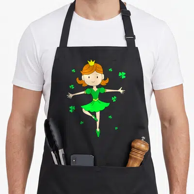 Dancing princess apron - Cartoon girl in green dress yellow crown surrounded by green shamrocks — shown on a black cotton canvas apron against a white background