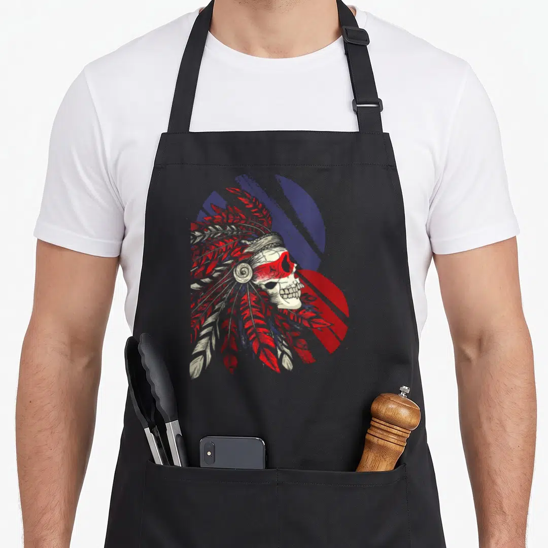 Skull native american aprons - Distressed skull in feathered headdress with red and blue circular background — shown on a black cotton canvas apron against a white background