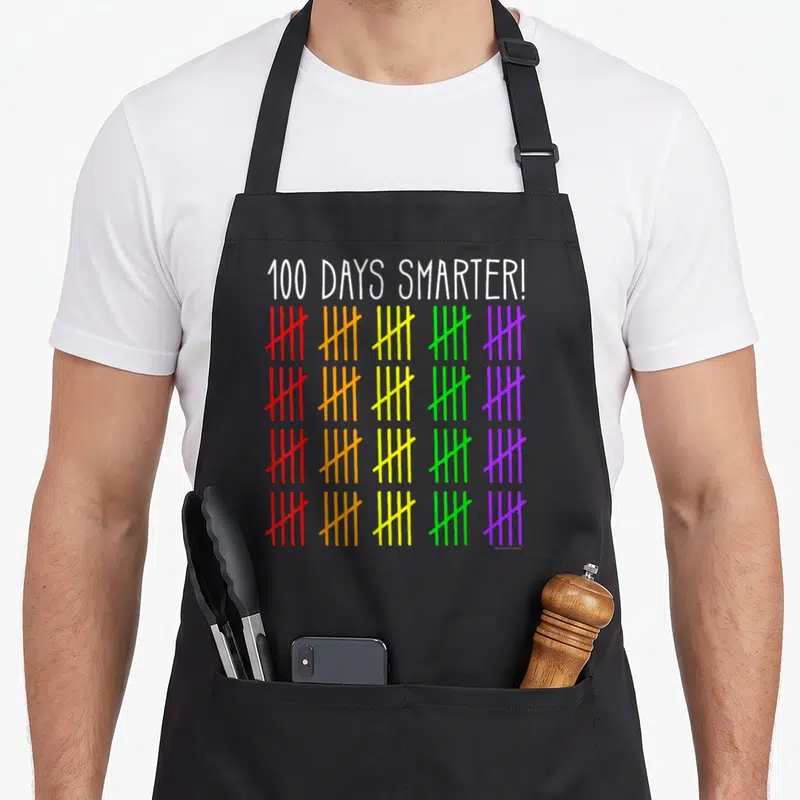 Smarter counting teacher apron - 100 DAYS SMARTER text above four rows of five colorful tally mark groups in red orange — shown on a black cotton canvas apron against a white background