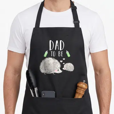 Shower animal apron - DAD TO BE text with two hedgehogs and green bottles — shown on a black cotton canvas apron against a white background