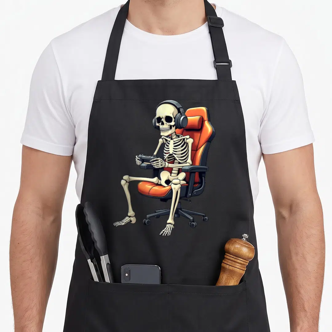 Skeleton gamer halloween apron - Cartoon skeleton sitting in orange gaming chair wearing headphones and holding — shown on a black cotton canvas apron against a white background