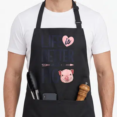Better pig farmers apron - Stacked purple text reading Life is Better with a Pig next to a pink cartoon pig head — shown on a black cotton canvas apron against a white background
