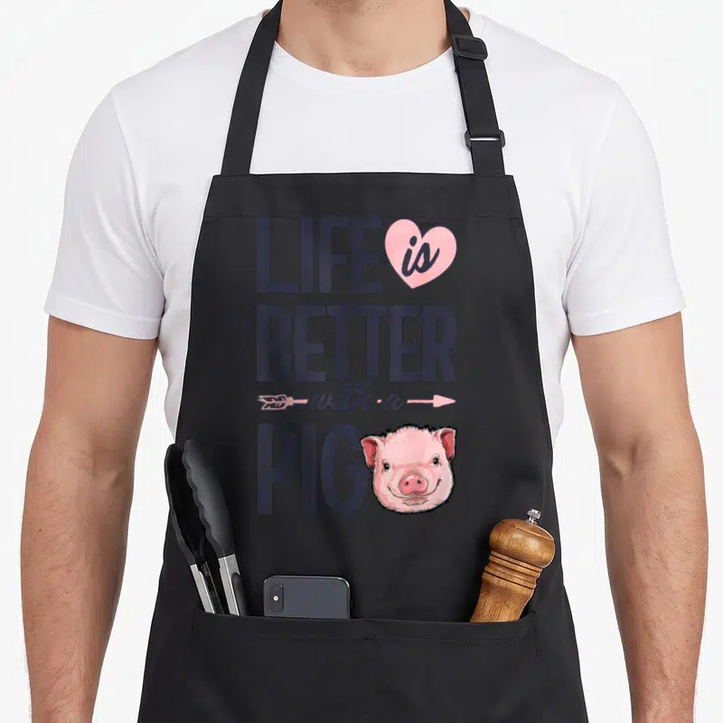 Better pig farmers apron - Stacked purple text reading Life is Better with a Pig next to a pink cartoon pig head — shown on a black cotton canvas apron against a white background