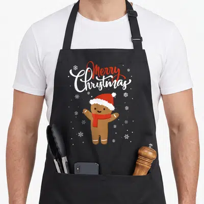 Merry baker apron - Merry Christmas text above a gingerbread man with hat and scarf amidst snowflakes — shown on a black cotton canvas apron against a white background