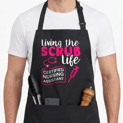 Living nursing aprons - Pink and white living scrub life text with stethoscope and syringe on black background — shown on a black cotton canvas apron against a white background