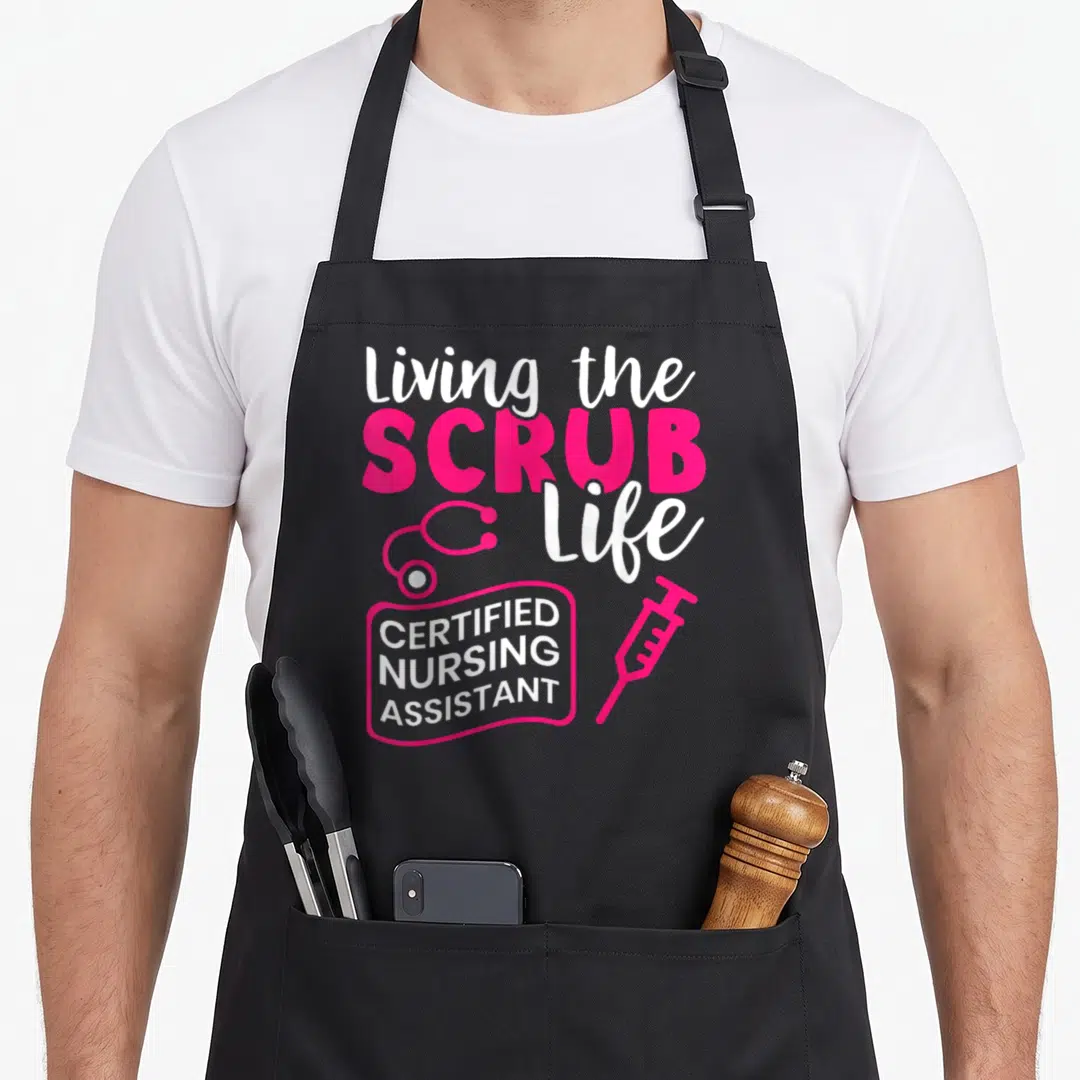 Living nursing aprons - Pink and white living scrub life text with stethoscope and syringe on black background — shown on a black cotton canvas apron against a white background
