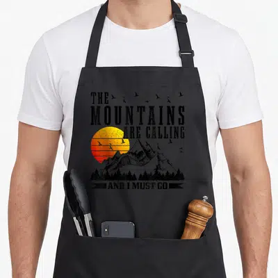 Mountain mountaineering apron - Distressed text and sun over mountains with birds and trees in yellow orange red — shown on a black cotton canvas apron against a white background