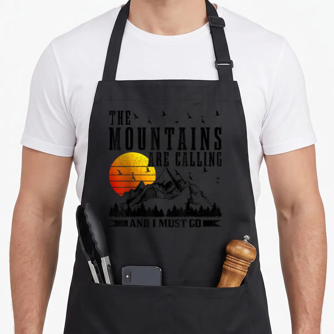 Mountain mountaineering apron - Distressed text and sun over mountains with birds and trees in yellow orange red — shown on a black cotton canvas apron against a white background
